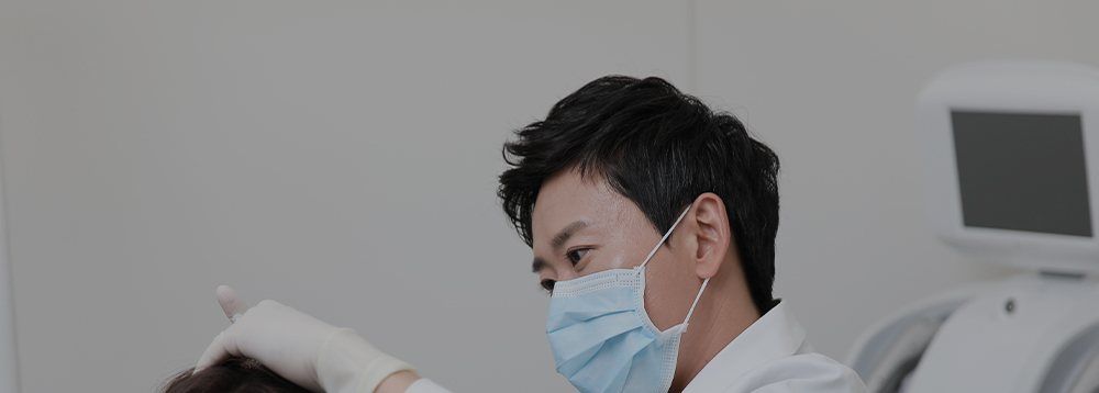 마스크 쓴 흰 가운 의사가 진료 의자에 앉은 환자의 머리를 짚으며 살핌